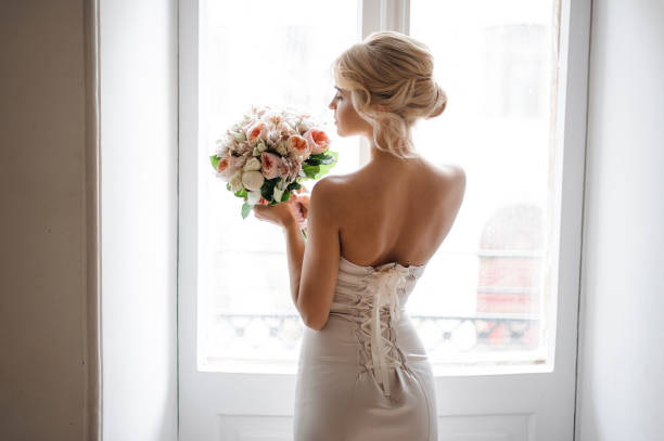 Back view of the elegant blonde bride dressed in a white dress holding a wedding bouquet on the background of window. Wedding concept