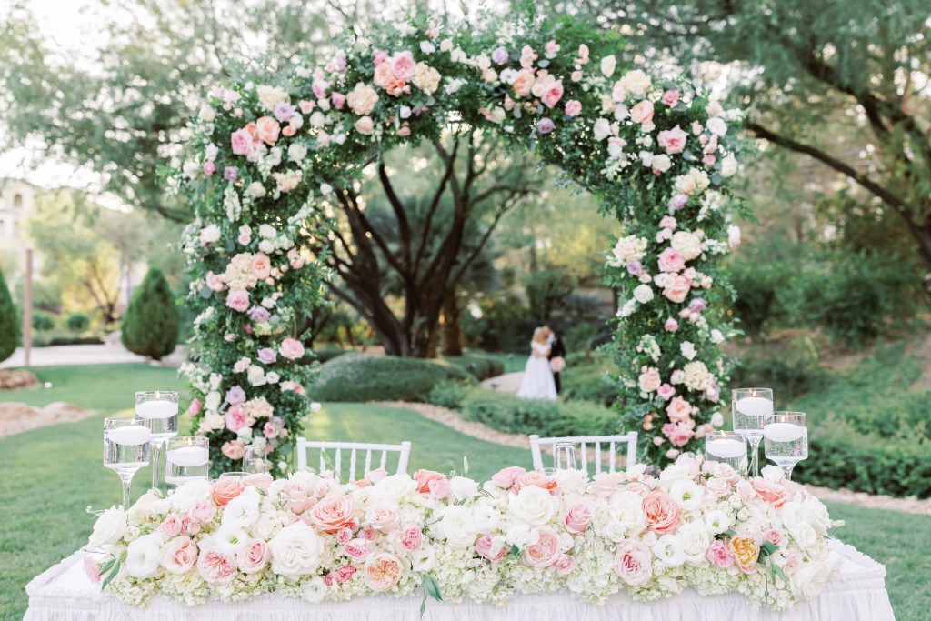 pink and white florals on sweetheart table with lush arbor overhead. Bride and Groom stand lovingly in the background at their Las Vegas Wedding.