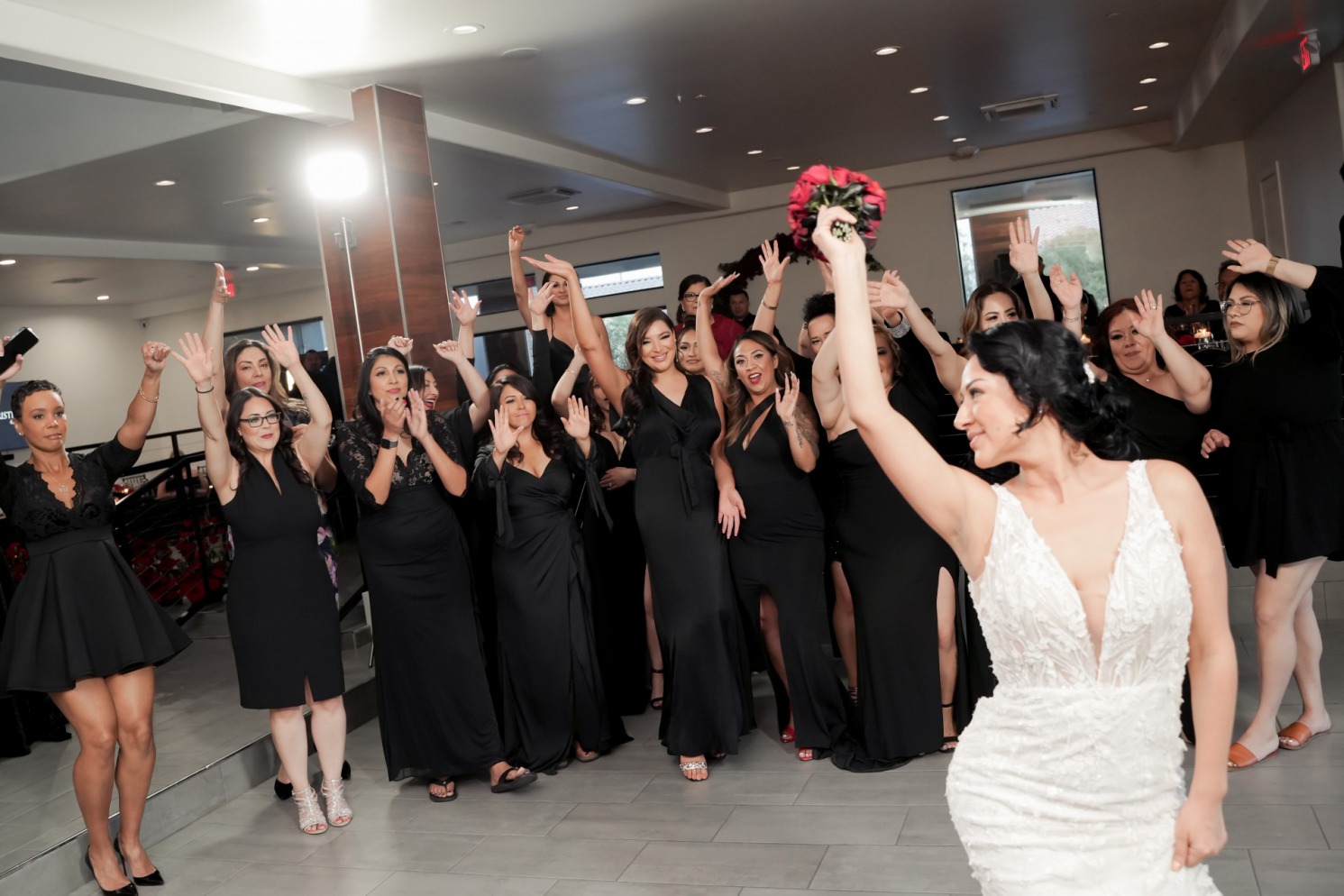 A Bride tosses the bouquet over her shoulder as her bridesmaids and guests dressed in black prepare to catch it