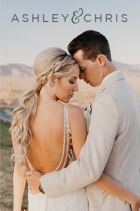 Ashley and Chris's romantic desert wedding. The couple poses in their wedding attire. She wears a low back dress with her hair hanging in a bohemian curled pony tail and he wears a tan suit in front of the stunning mountain views