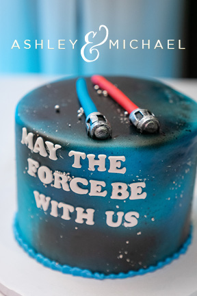 Nerdy Star Wars wedding cake. The background looks like the galaxies with the words "May the force be with us" A blue and red light saber sit atop the cake by manon bakery
