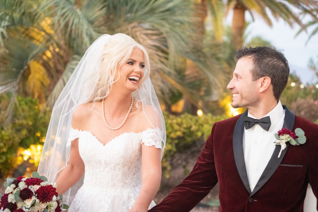 Beautiful bride with a curly blonde updo and veil wearing a sweetheart gown with off the shoulder sleeves holds hands with her new husband wearing a burgundy velvet shawl tuxedo at an outdoor Las Vegas Wedding Ceremony.