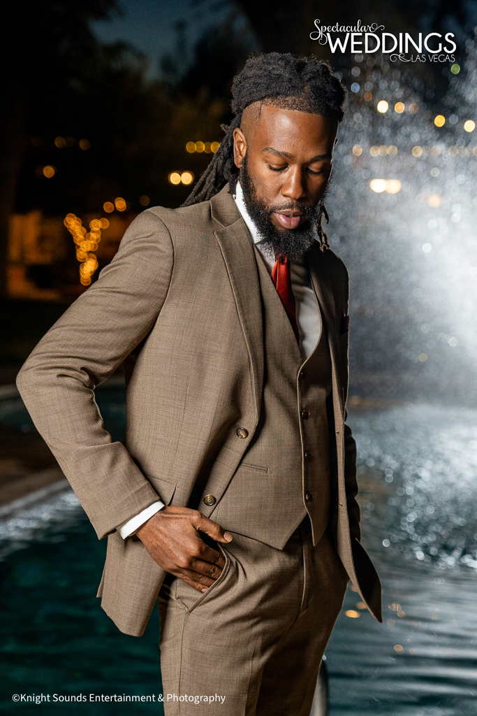 Handsome African American Groom wearing tan suit with orange accessories poses in front of a dramatic fountain at his wedding.