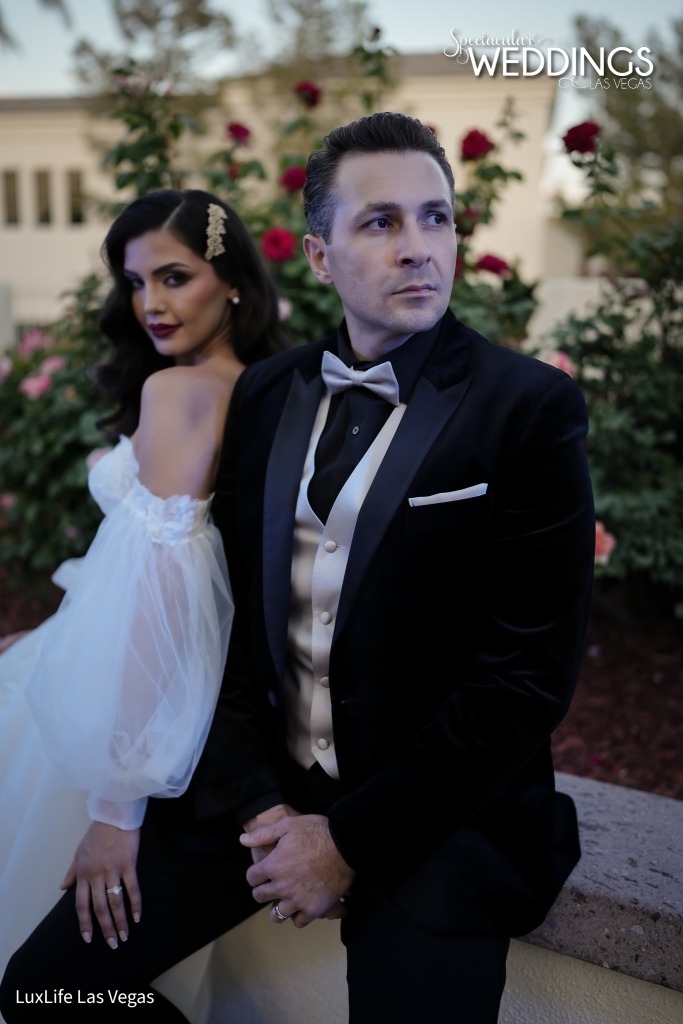 Cassic black wedding tux with black shirt, gold vest and bowtie from Friar Tux. Groom sits in his wedding tuxedo next to his lovely bride with a romantic wedding gown